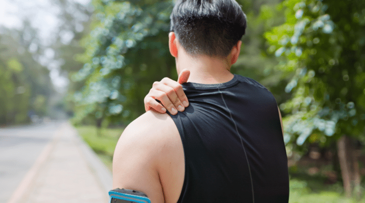 closeup back view asian young man is sporting in park he stand by roadside suffers from sprain back shoulder pain with hands massage the painful point