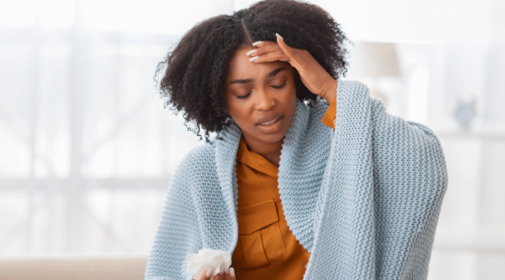 Distressed african american young woman with curly hair sitting on a couch, wrapped in a blue blanket, holding a tissue and touching her forehead
